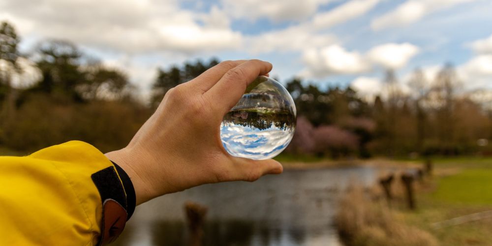 Ruka držiaca priehľadnú sklenenú guľu, ktorá zrkadlí jazero a okolitú prírodu pod oblačnou oblohou. Symbolizuje detailný pohľad a dôležitosť ochrany vodných zdrojov a legislatívnych povinností v oblasti ochrany vôd pre podnikových ekológov.